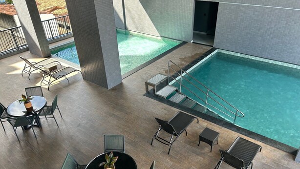 An indoor pool area with tiled walls, a pool, and multiple seating areas with chairs and tables. - Bela Hospedagem - 105 Solaz Smart (João Pessoa)