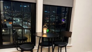A corner room with a city view, a round table, three black chairs, and a small candle on the table.