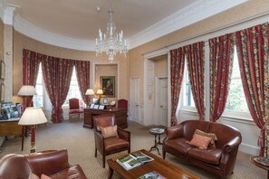 A room with a chandelier, leather armchairs, a desk with a lamp, and floral curtains. - Dukes Hotel (Bath)