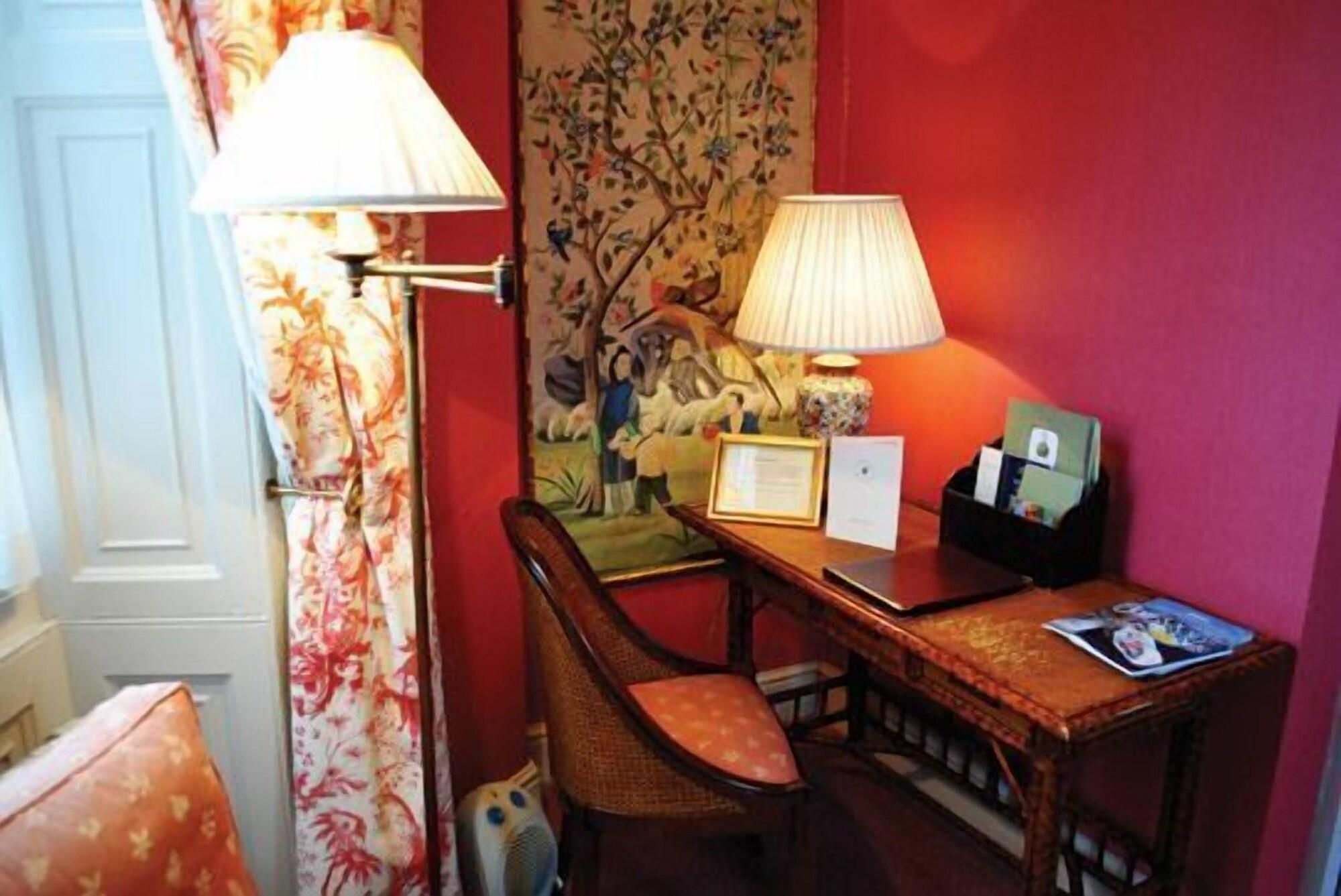 Una habitación con escritorio, silla, lámparas, un certificado enmarcado y una mesita con una revista.