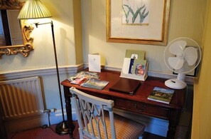 A hotel room with a wooden desk, a white chair, a radiator, a lamp, a framed picture, a fan, and a magazine. - Dukes Hotel (Bath)