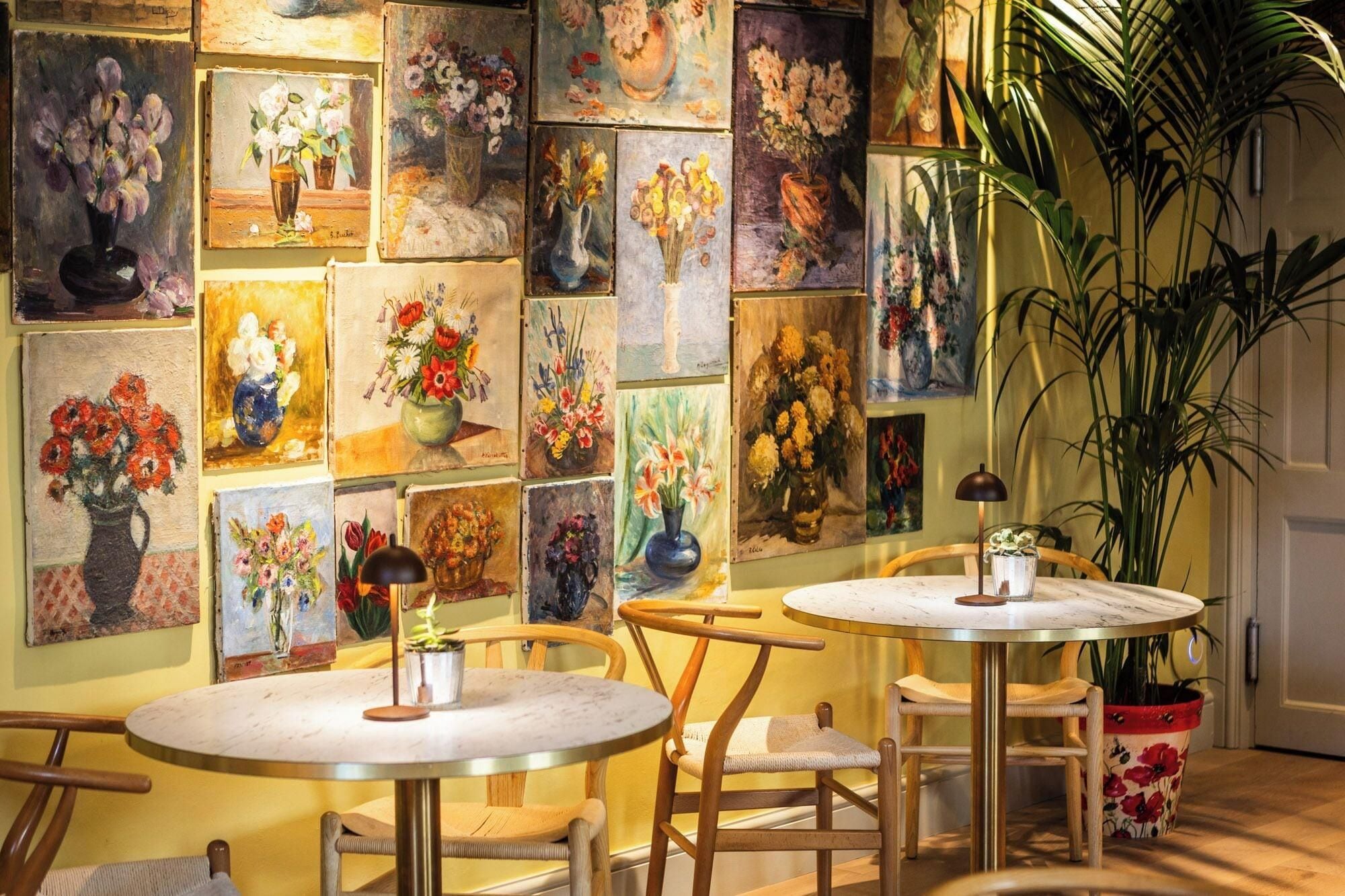 A room with multiple round tables and chairs, a large potted plant, and a wall adorned with various flower paintings.