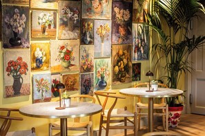 A room with multiple round tables and chairs, a large potted plant, and a wall adorned with various flower paintings. - Dukes Hotel (Bath)