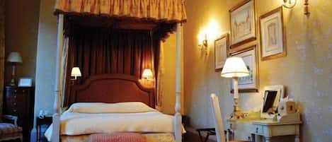 A hotel room with a four-poster bed, a desk with a chair, and a small table with a lamp.