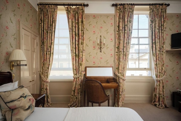 A hotel room with floral wallpaper, a bed, a desk with a chair, a lamp, and a television.