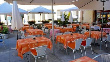 Ein Straßencafé mit Tischen und Stühlen, beschattet von Sonnenschirmen, mit einem Gebäude im Hintergrund.
