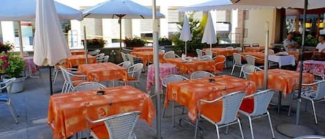 Um café ao ar livre com mesas e cadeiras, coberto por guarda-sóis, e um edifício ao fundo.