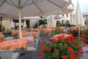 Un área de estar al aire libre con mesas y sillas bajo sombrillas, rodeada de plantas y flores en macetas.