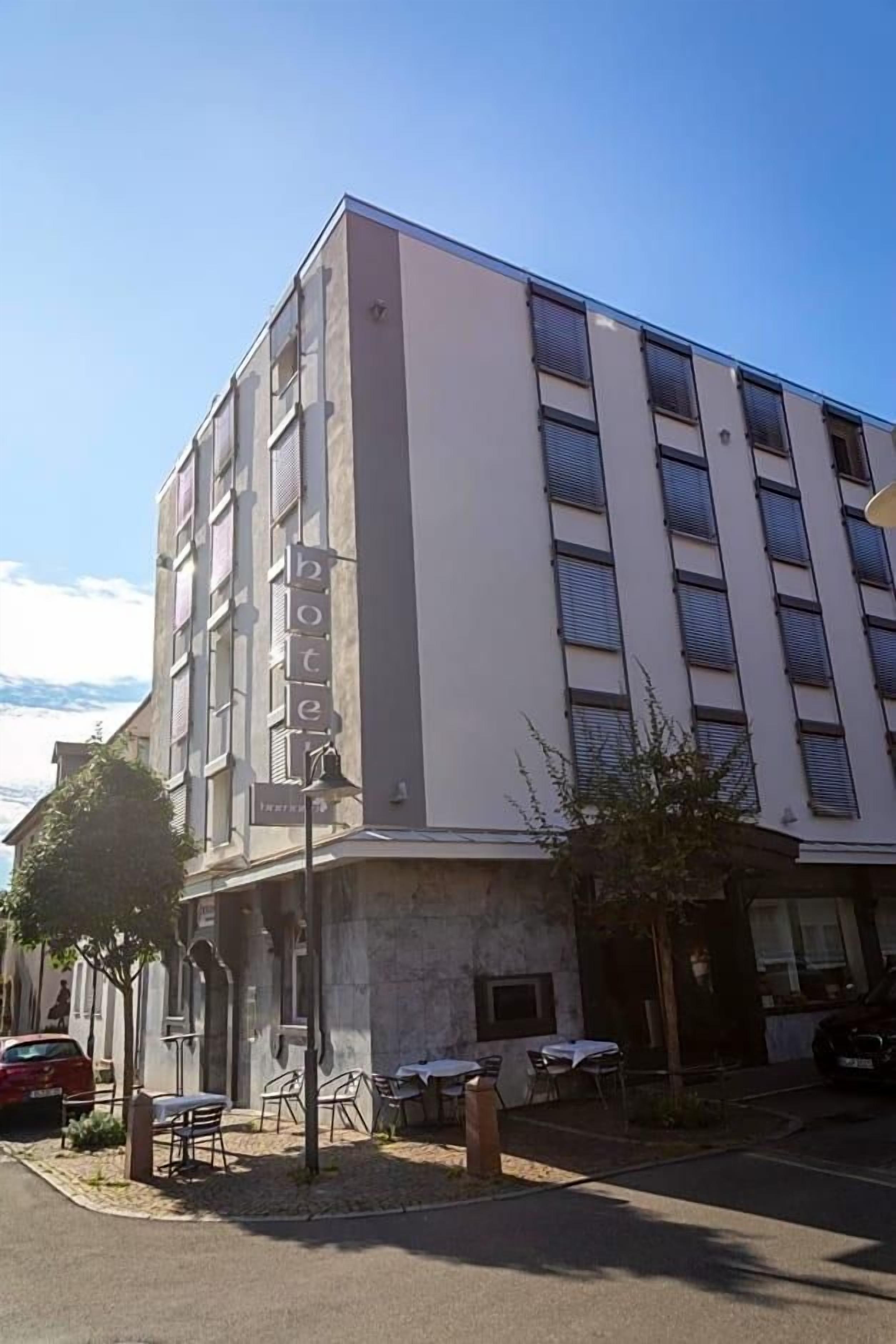 Un hotel de varios pisos con un llamativo letrero de 'hotel' en la fachada, mesas al aire libre y un área de estacionamiento.