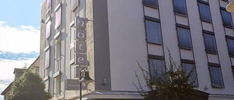 Un hotel de varios pisos con un llamativo letrero de 'hotel' en la fachada, mesas al aire libre y un área de estacionamiento.