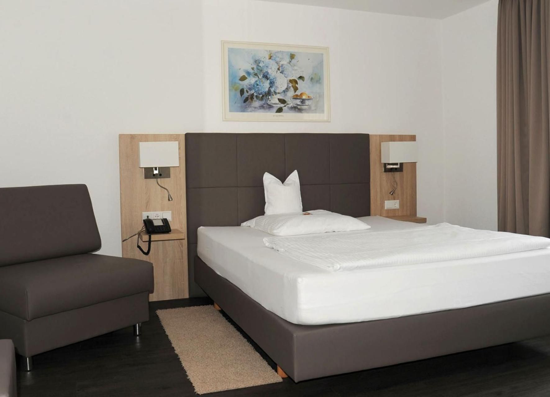 A hotel room with a bed, a sofa, a bedside table with a lamp, a phone, and a painting on the wall.