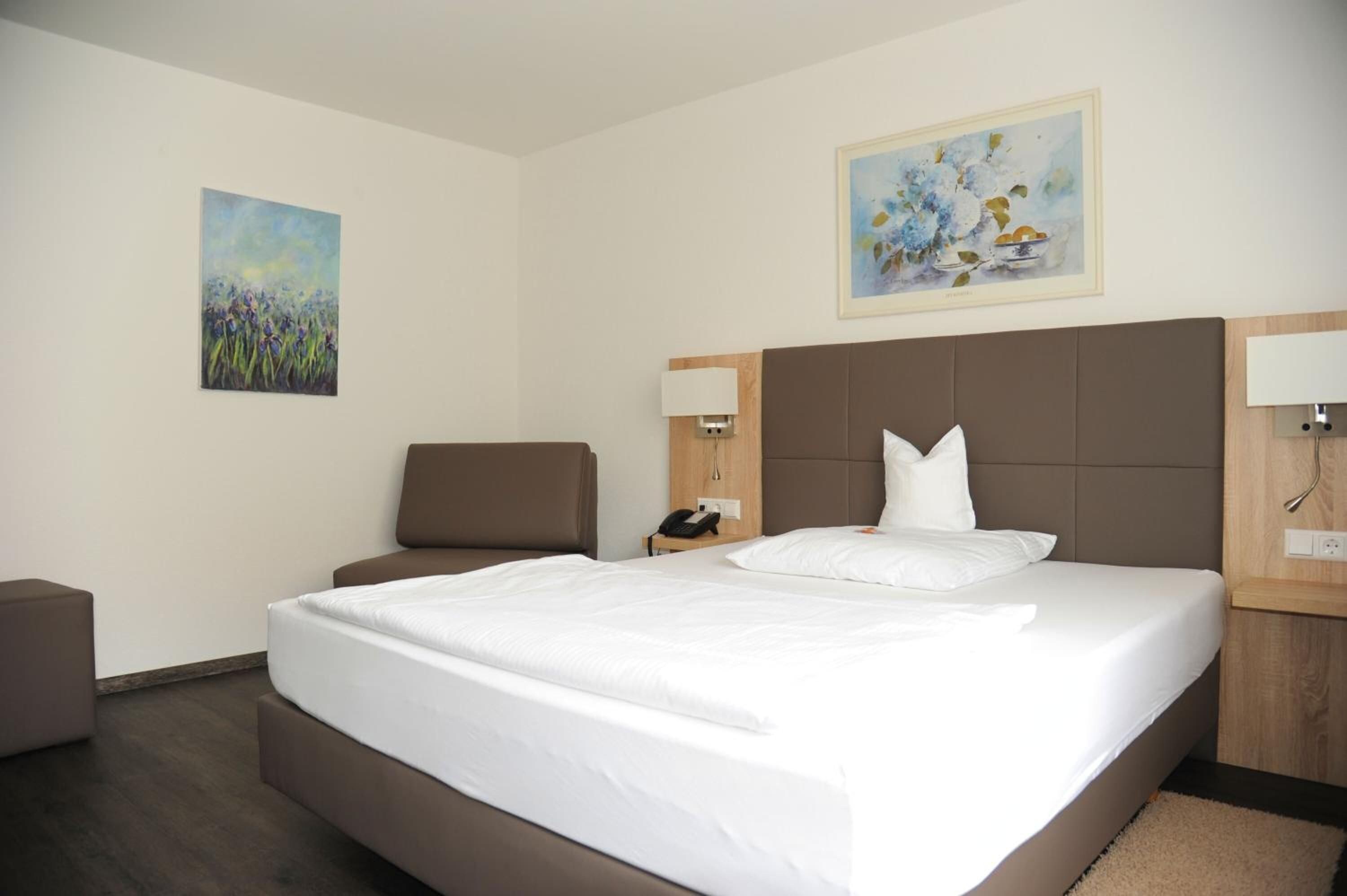 A hotel room with a large bed, a chair, a desk, and two paintings on the walls.