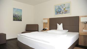A hotel room with a large bed, a chair, a desk, and two paintings on the walls.