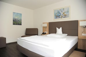 Ett hotellrum med en stor säng, en stol, ett skrivbord och två tavlor på väggarna.