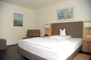 A hotel room with a large bed, a bedside table with a lamp, a chair, and two paintings on the walls. - Hamann (Balingen)