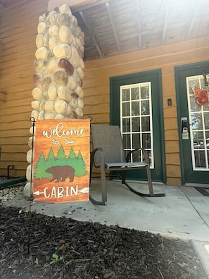 A cabin entrance with a welcome sign, a stone pillar, a rocking chair, and a door with a window.