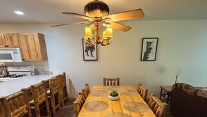 Un comedor con mesa y sillas de madera, un ventilador de techo y una cocina al fondo.