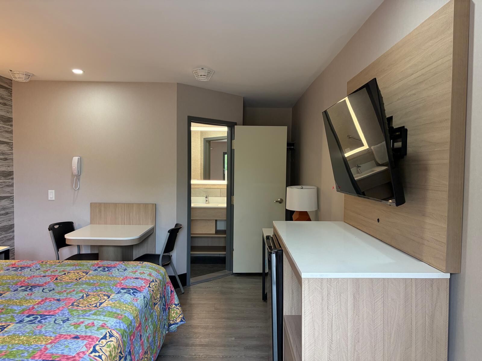 A hotel room with a bed, a desk, a chair, a television mounted on the wall, and a bathroom visible through an open door.