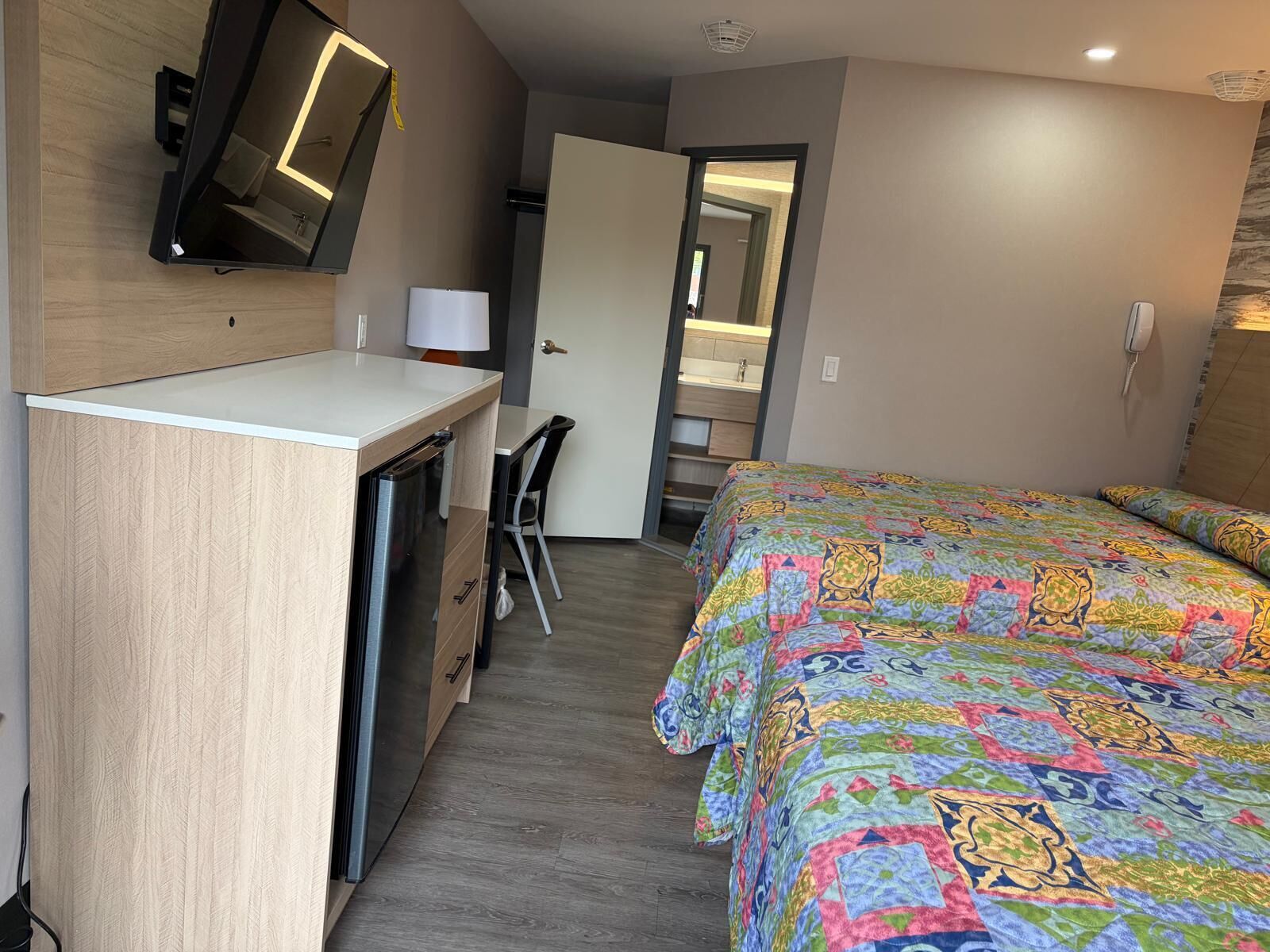 A compact hotel room with a bed, a desk, a chair, a mirror, and a television.