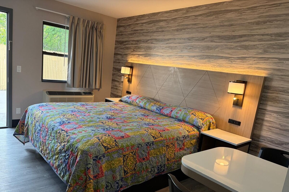 Una habitación de hotel con una cama, mesitas de noche, un escritorio y un ventanal con cortinas.
