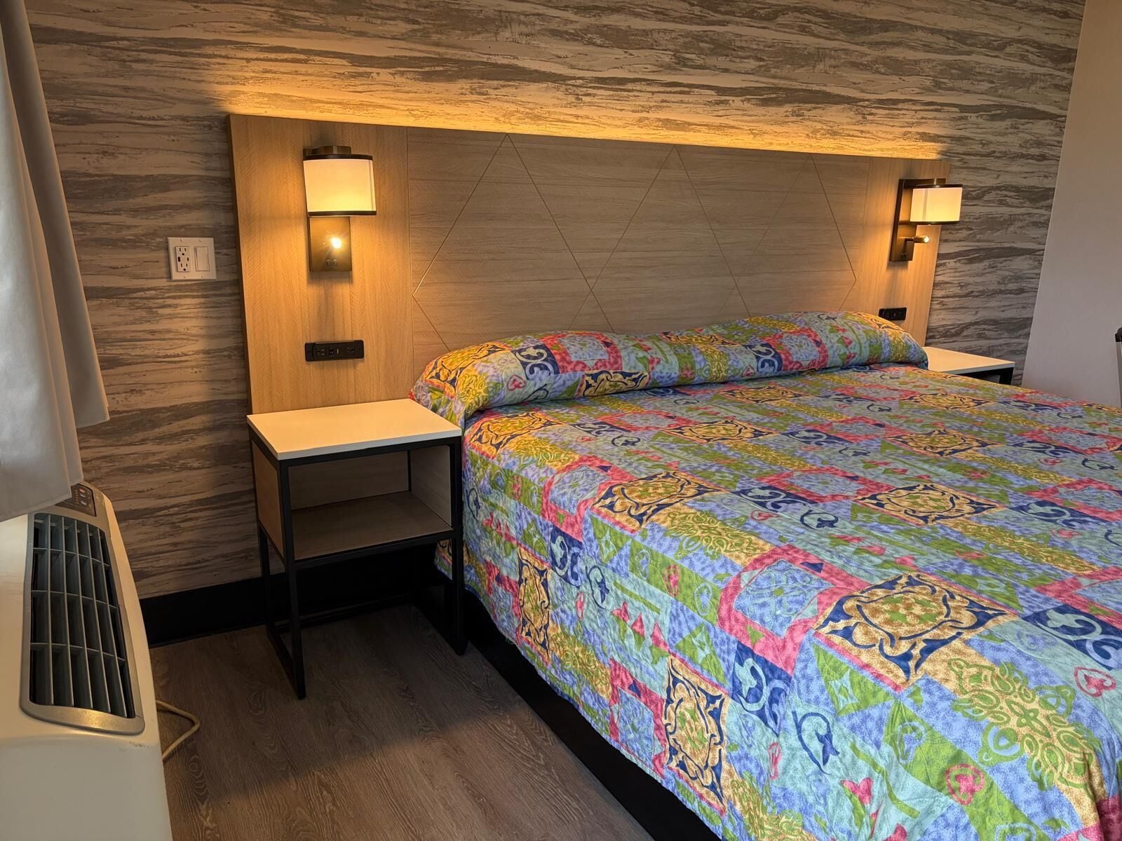 A bedroom with a patterned bedspread, two bedside lamps, a wooden headboard, and a small nightstand.