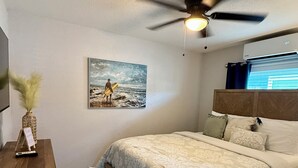 Een slaapkamer met een bed, een plafondventilator, een schilderij, een nachtkastje en een raam met gordijnen.