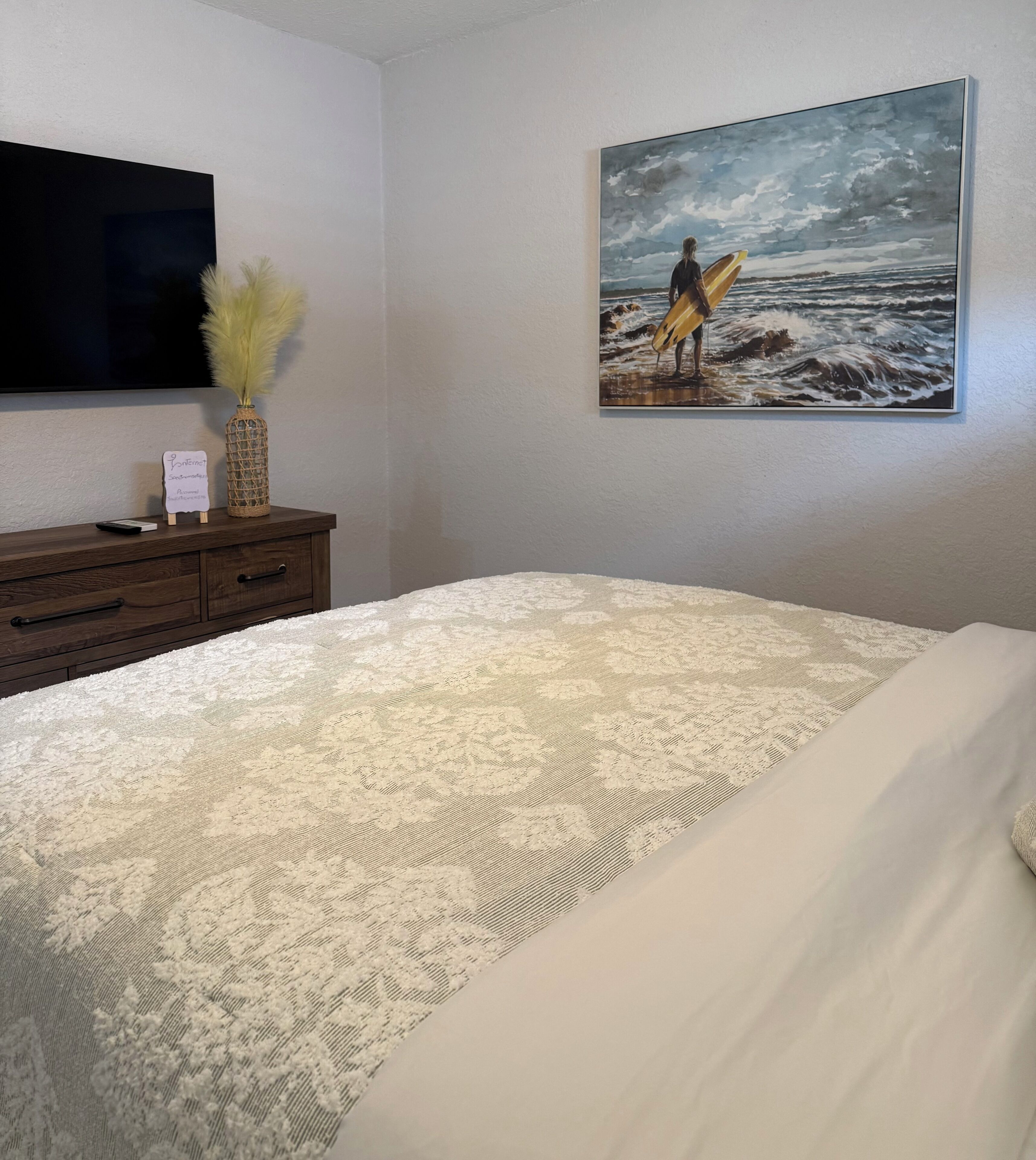 A bedroom with a bed, a dresser, a painting of a surfer, and a flat-screen TV.