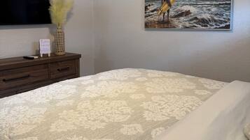 A bedroom with a bed, a dresser, a painting of a surfer, and a flat-screen TV.