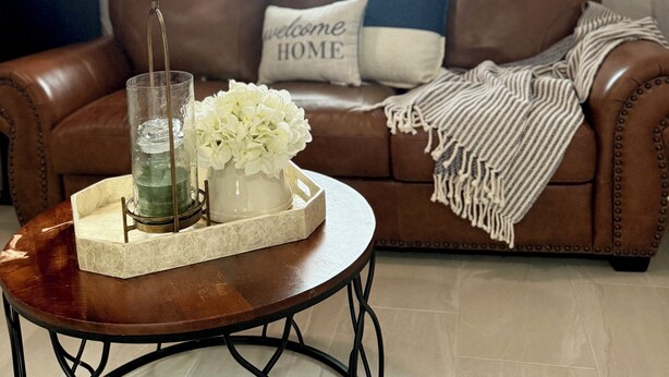 Een bruine leren bank met een 'welcome home' kussen, een glazen kandelaar met bloemen en een gestreept plaid.