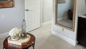 Een hal met een witte deur, een ronde tafel met een vaas en een kaars, en een grote spiegel aan de muur.