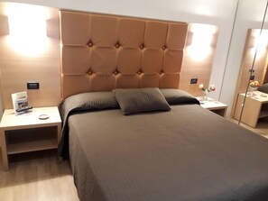 Una habitació d'hotel moderna amb un capçal de llit amb textura, un llit gran, dues taules de nit i una petita taula auxiliar amb un comandament a distància.