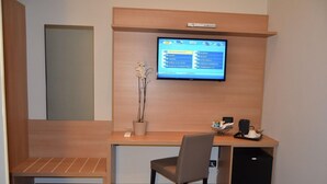 A hotel room with a wooden desk, a chair, a television mounted on the wall, a potted plant, and a small cabinet.