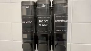 Trois distributeurs noirs, portant les inscriptions « SHAMPOO », « GEL DOUCHE » et « APRÈS-SHAMPOO », sont fixés à un mur recouvert de carreaux blancs.