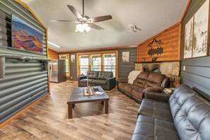 Ein Interieur mit Holzbalkendecke, Deckenventilator, Kamin, Sofa, Couchtisch und Kühlschrank.