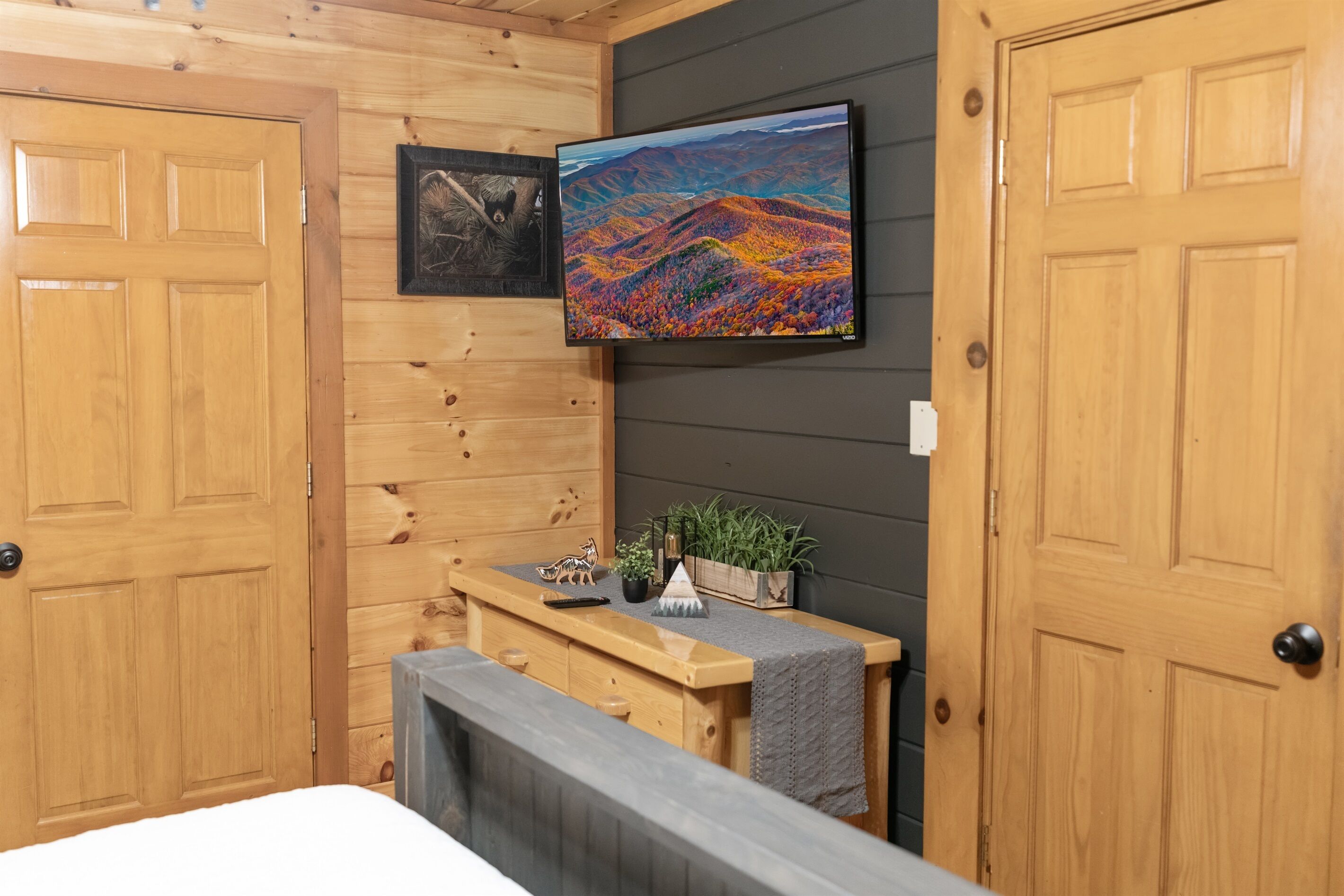 A wooden cabin interior with a bed, a wooden bench, a mounted painting, and a small table with a plant and a figurine.