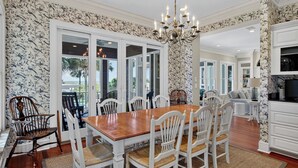 A dining area with a wooden table and white chairs, a chandelier, and a view through glass doors. - 3902 Palm Blvd by Avantstay Beach Access! Oceanfront Views w/ Pool (Isle of Palms)