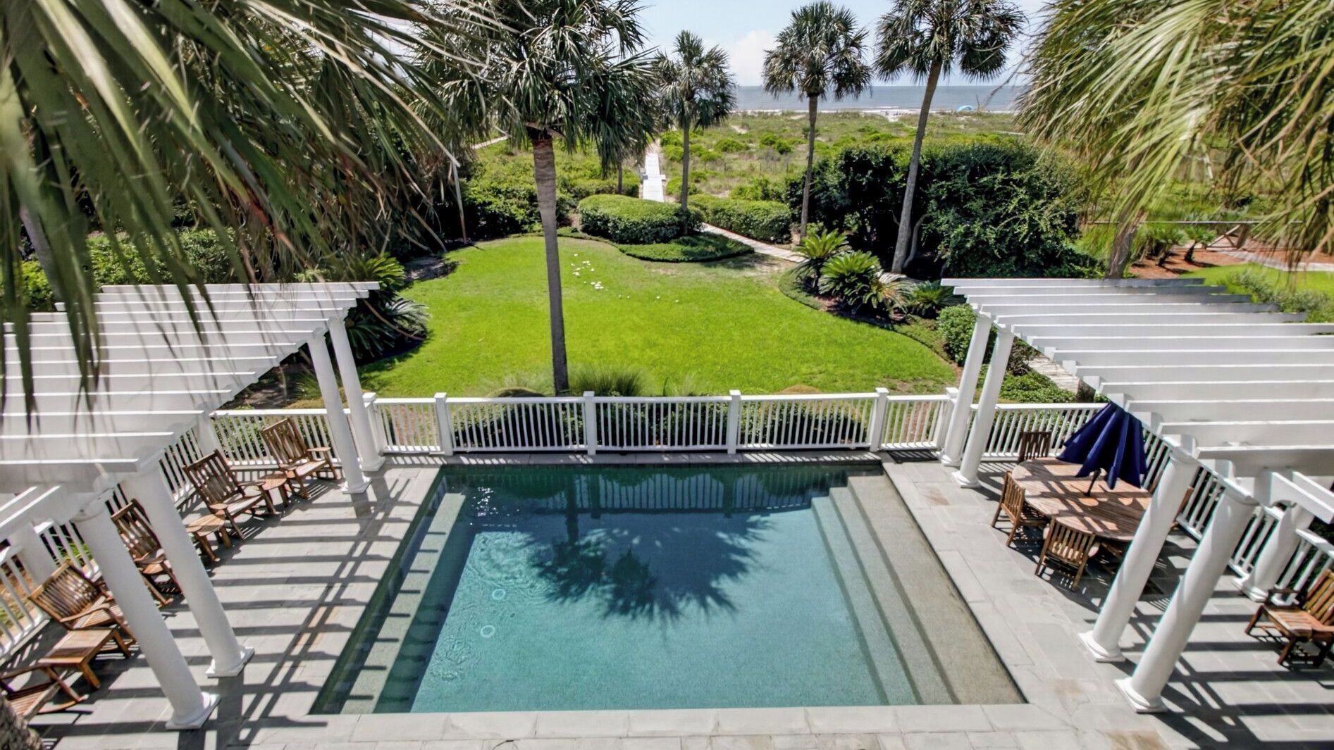 A pool with a white deck and chairs, surrounded by palm trees and a view of the ocean.