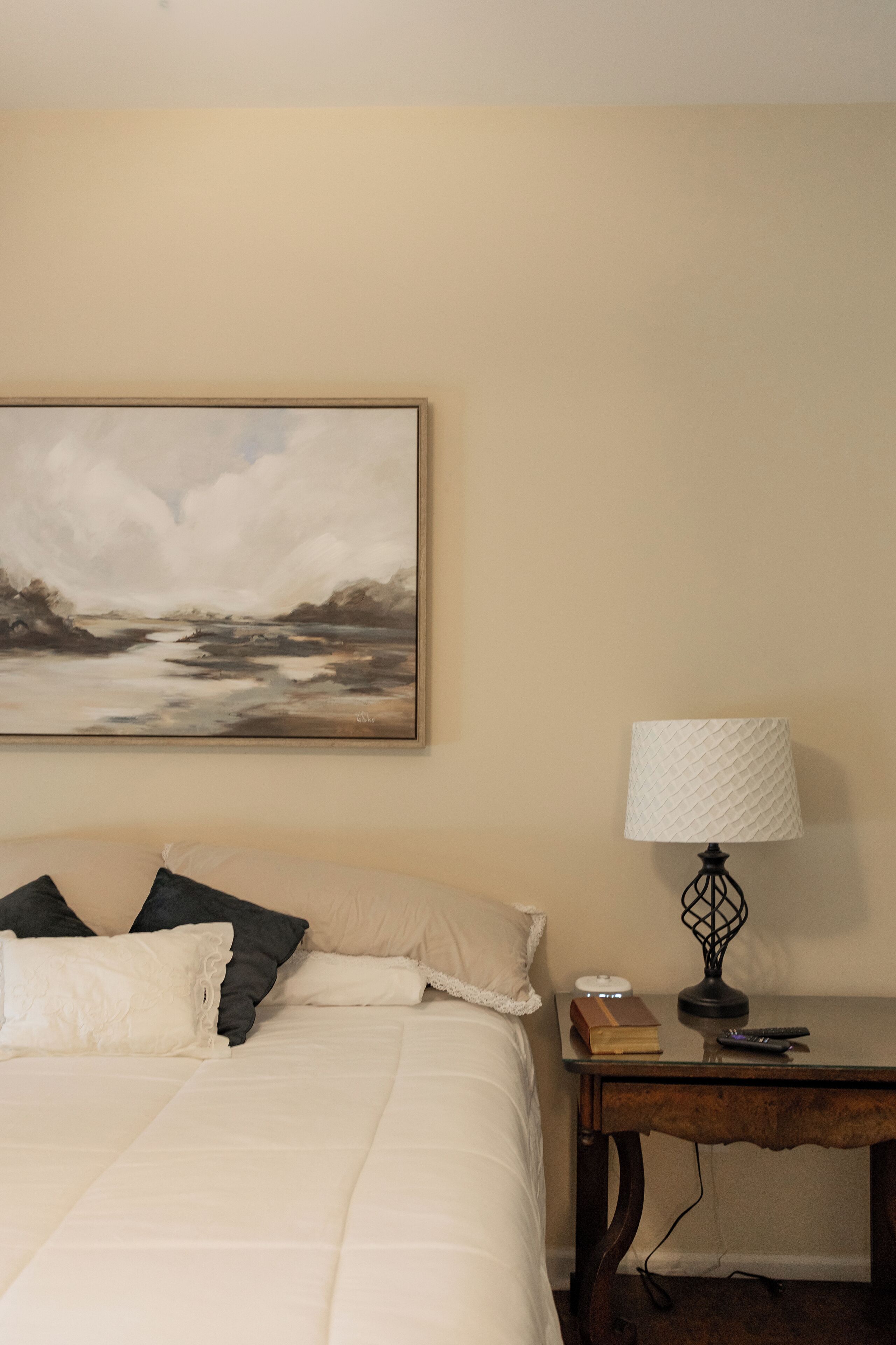 Une chambre à coucher avec un lit, des oreillers, une table de chevet avec une lampe, un livre et un tableau accroché au mur.