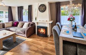 Een knusse woonkamer met een open haard, een eethoek met een kroonluchter en een groot raam met gordijnen.