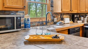 A kitchen with wooden cabinets, a stone backsplash, a window with a view of trees, a microwave, a sink, and a countertop with a tray of snacks. - Turkey Run, Cozy Woodland Cabin Retreat in the Heart of Hocking Hills, Hot Tub (Logan)