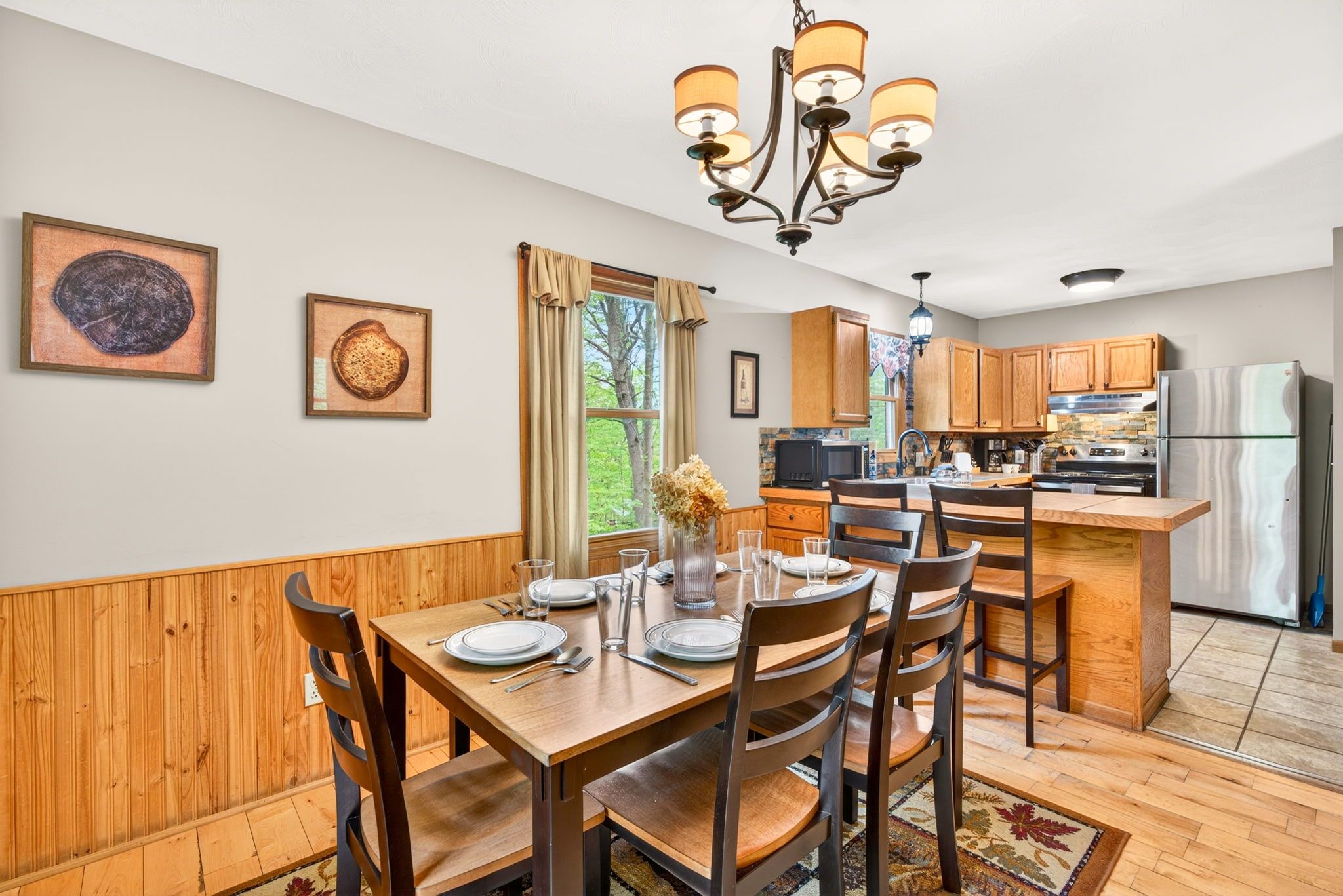 A dining area with a wooden table and chairs, a chandelier, and a kitchen in the background.
