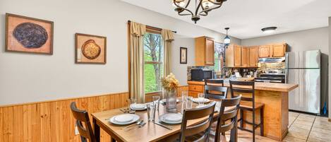 A dining area with a wooden table and chairs, a chandelier, and a kitchen in the background.