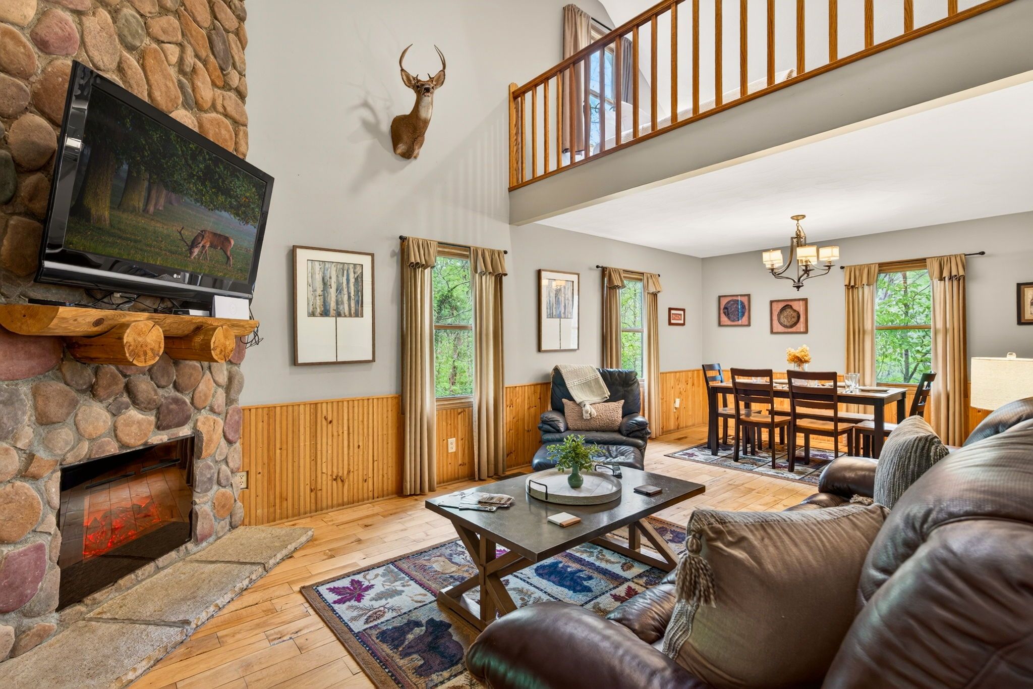A spacious living room with a stone fireplace, a mounted deer head, a television, and a dining area with a chandelier.