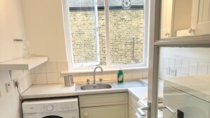 A compact kitchen with a washing machine, a sink, and a window with a view of a brick wall. - Charming London Apartment in Chelsea/ Fulham (London)