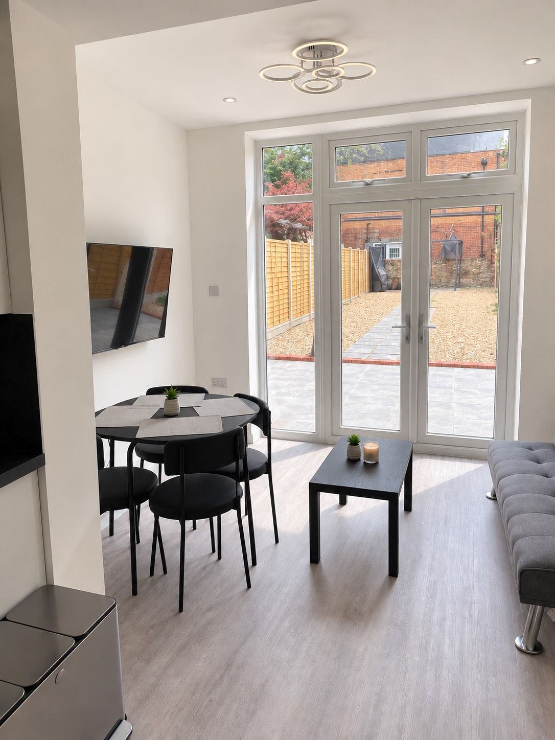 Un salon moderne comprenant un coin repas, un canapé gris, un téléviseur fixé au mur et une porte-fenêtre coulissante donnant sur un jardin.