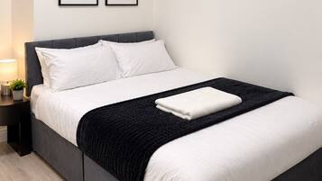 A modern bedroom with a dark grey bed, white bedding, and a black throw blanket. There are two framed abstract artworks on the wall above the bed.
