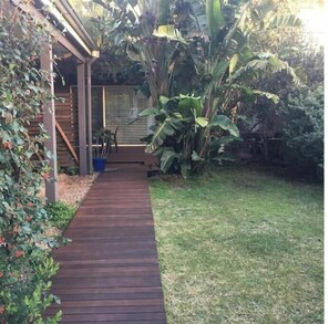 Ein Holzweg führt zu einem Gebäude mit überdachter Terrasse, umgeben von üppiger Vegetation und Bananenpflanzen.