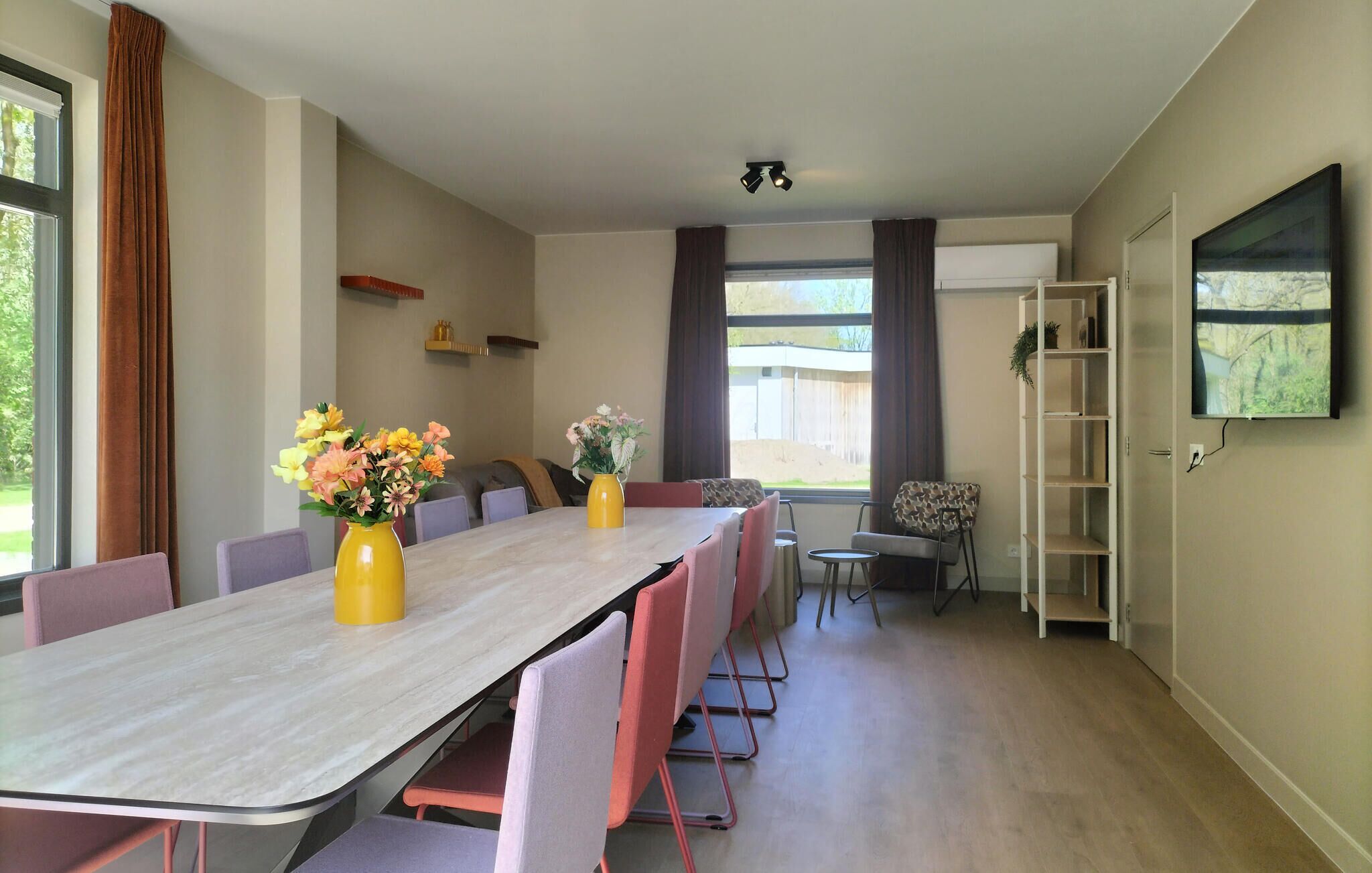 A dining area with a long table, chairs, vases with flowers, a TV mounted on the wall, and a view of the outdoors.