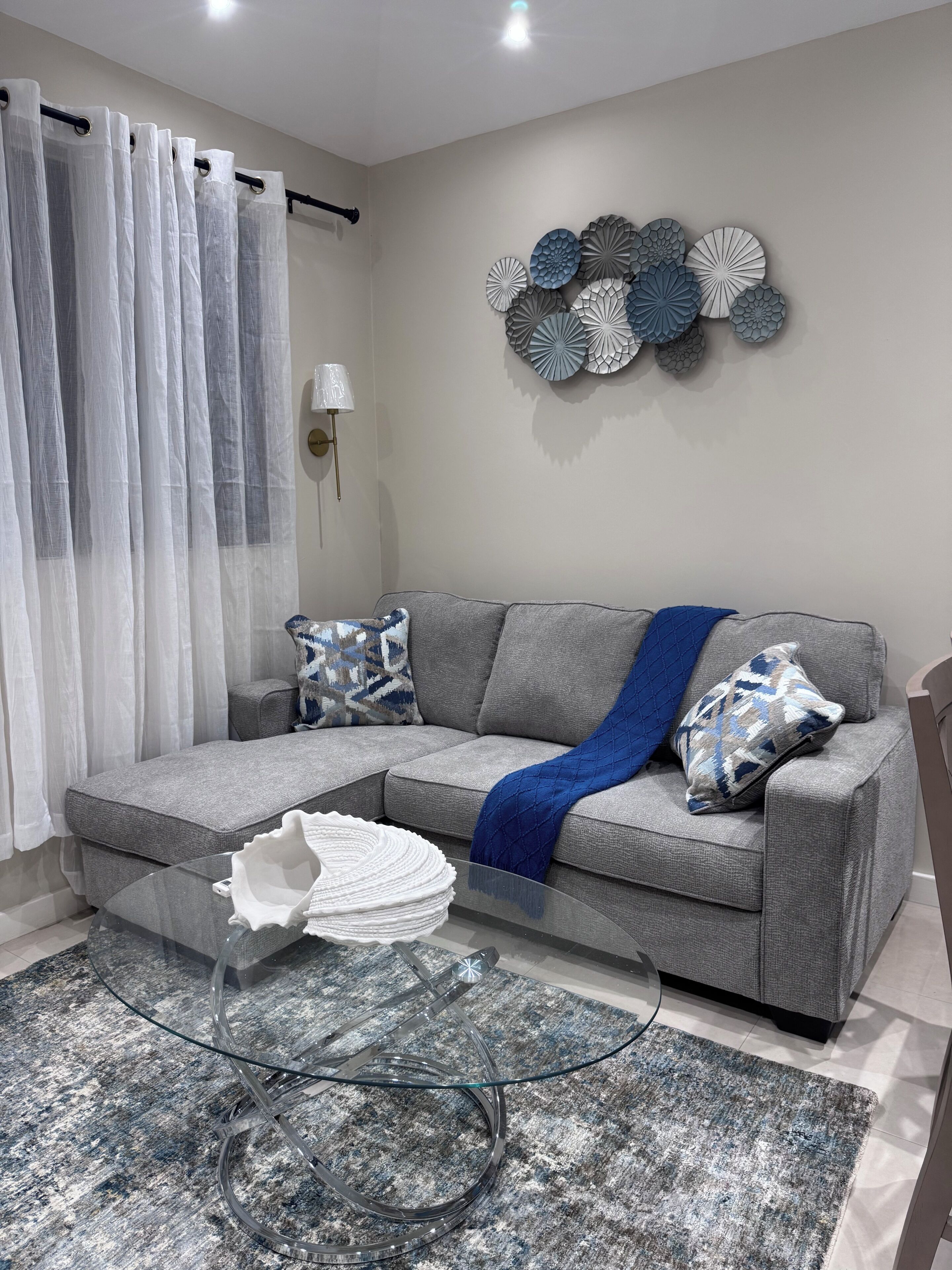 A living room with a grey sofa, patterned cushions, a glass coffee table, a decorative wall piece, and a curtain.