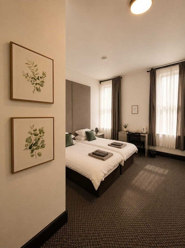 Une chambre d’hôtel avec un lit double, un bureau, deux reproductions botaniques encadrées accrochées au mur et un sol recouvert de moquette.